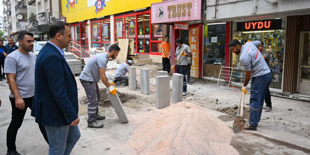 Bornova Zafer Caddesi'nde belediye ekiplerinin yürüttüğü kaldırım düzenleme ve çevre düzenlemesi çalışmaları sırasında, üzerinde Bornova Belediyesi logolu gri tişörtler bulunan işçiler beton blokları yerleştirip kürekle kum seriyor. Çalışmaları izleyen iki erkek yetkili de işçilerin yanında duruyor; arka planda ise Şok market ve çeşitli dükkanların bulunduğu cadde üzerindeki hareketlilik görülüyor.