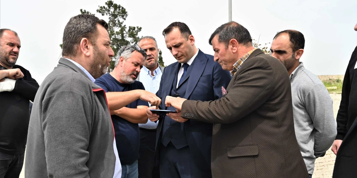 Lacivert takım elbiseli bir erkek, çevresindeki bir grup erkekle birlikte dış mekanda elindeki tableti veya telefonu inceliyor. Grubun içindeki diğer kişiler de merakla ekrana bakarak fikir alışverişinde bulunuyor.