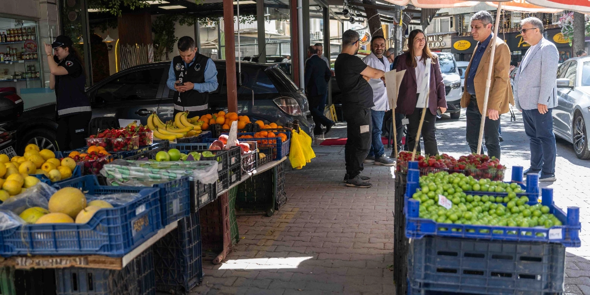 Bornova'da bir manav tezgahının önünde denetim yapan zabıta ekipleri ve yetkililer yer alıyor. Mavi plastik kasalar içinde sergilenen limon, çilek, muz, portakal ve yeşil erik gibi taze meyvelerin bulunduğu tezgahın başında, üzerinde 