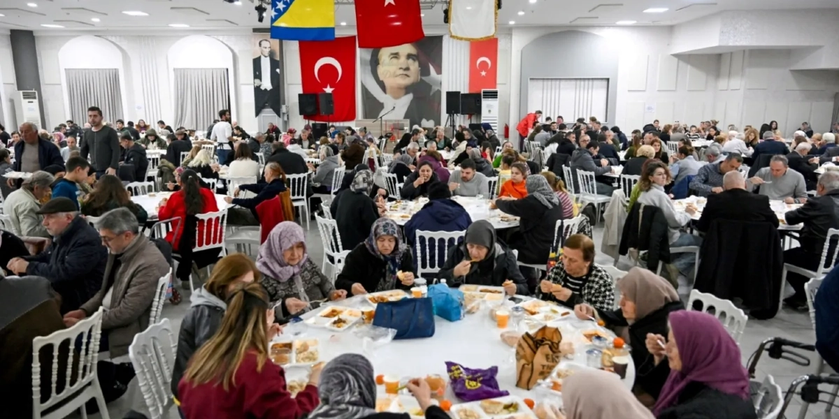 Geniş bir düğün veya etkinlik salonunda, yuvarlak masaların etrafında toplanmış kalabalık bir grup insan birlikte yemek yiyor. Arka planda duvarda büyük bir Mustafa Kemal Atatürk posteri, Türk bayrakları ve Bosna-Hersek bayrağı asılı. Ön plandaki masada başörtülü ve başı açık kadınlar, önlerindeki tabaklardan yemeklerini yiyor; salonun genelinde ise farklı yaş gruplarından insanlar sohbet ederek yemeklerini yiyor.