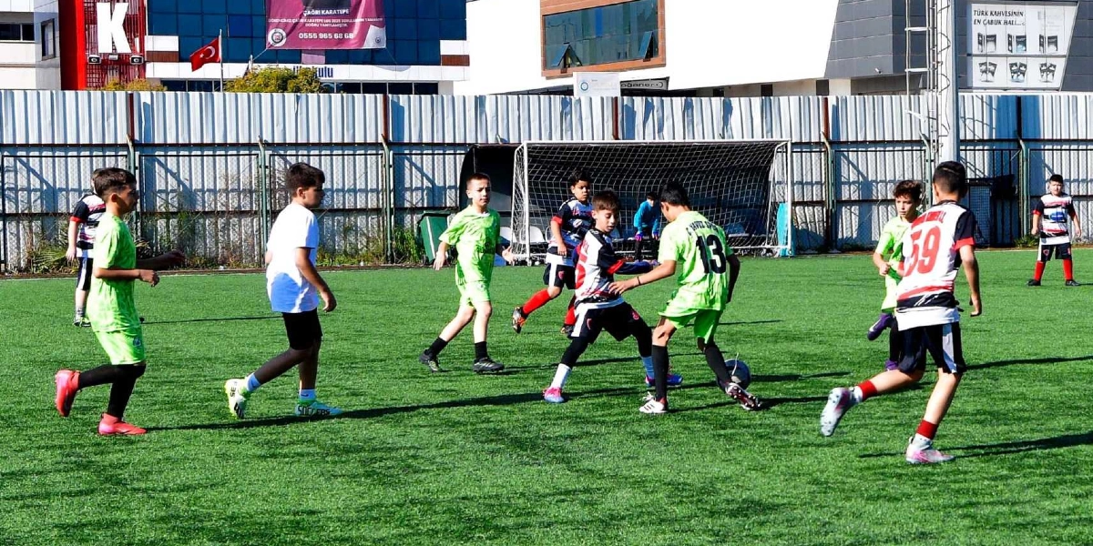Açık havada, suni çim kaplı bir sahada futbol maçı yapan çocuklar. Farklı takımlardan oyuncular, yeşil ve beyaz formalarla top sürme ve paslaşma mücadelesi veriyor. Arka planda metal çitler, bir kale direği ve binalar yer alıyor.