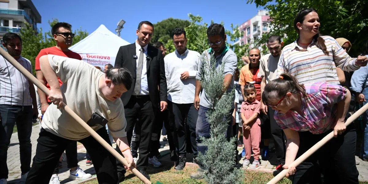 Açık havada, güneşli bir günde düzenlenen fidan dikme etkinliğinde, Down sendromlu iki genç ellerinde küreklerle yeni dikilen bir çam fidanının etrafındaki toprağı düzeltiyor. Gençlerin etrafında, aralarında Bornova Belediyesi logolu beyaz bir çadırın da bulunduğu alanda, onları izleyen ve destek veren çeşitli yaş gruplarından insanlar yer alıyor.