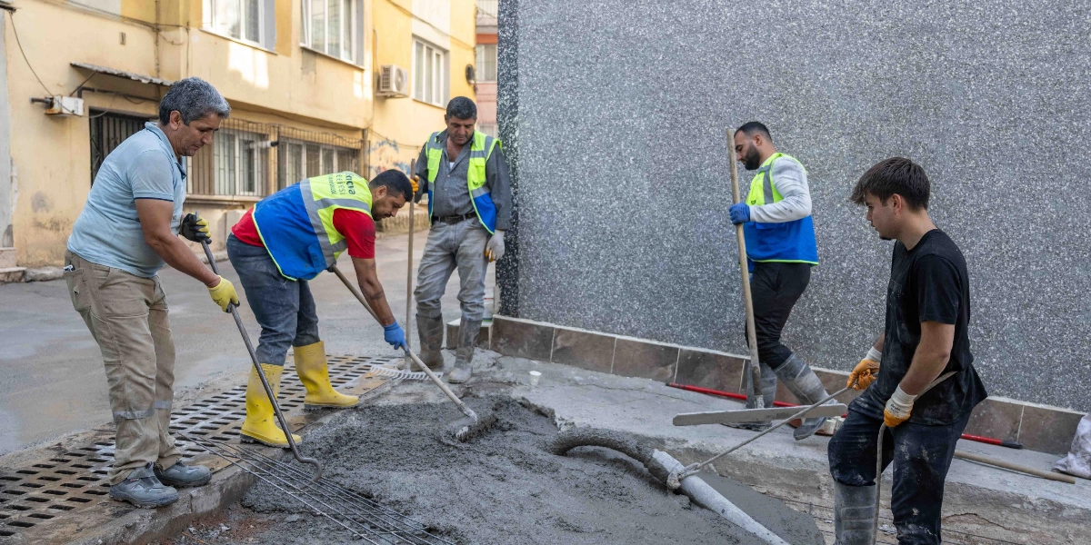 Bornova'da bir sokakta yol çalışması yapan beş kişilik işçi ekibi, taze dökülen betonu düzeltiyor ve tesviye ediyor. İşçilerden bazıları üzerinde Bornova Belediyesi yazılı reflektörlü yelekler ve sarı iş çizmeleri giyiyor, ellerinde mala, tırmık ve kürek gibi aletlerle beton yüzeyini pürüzsüzleştirmek için çalışıyor.