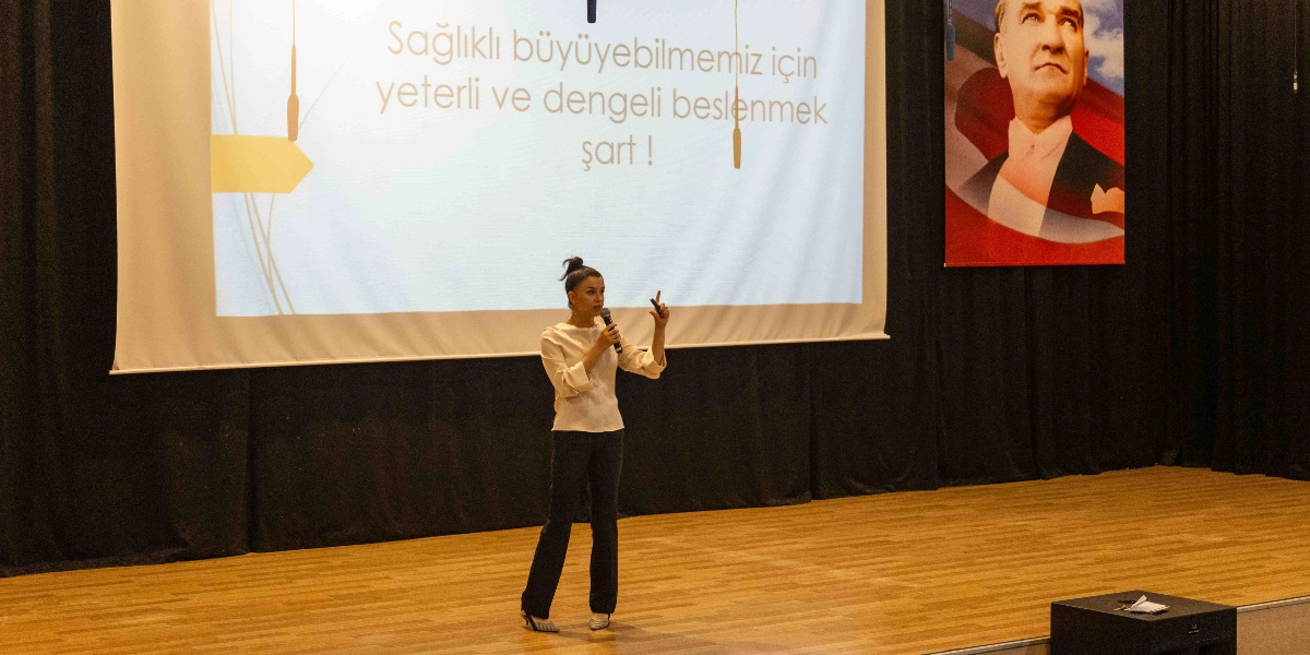 Konferans salonunda sahnenin ortasında duran bir kadın, elinde mikrofonla sunum yapıyor. Arkasındaki dev ekranda 