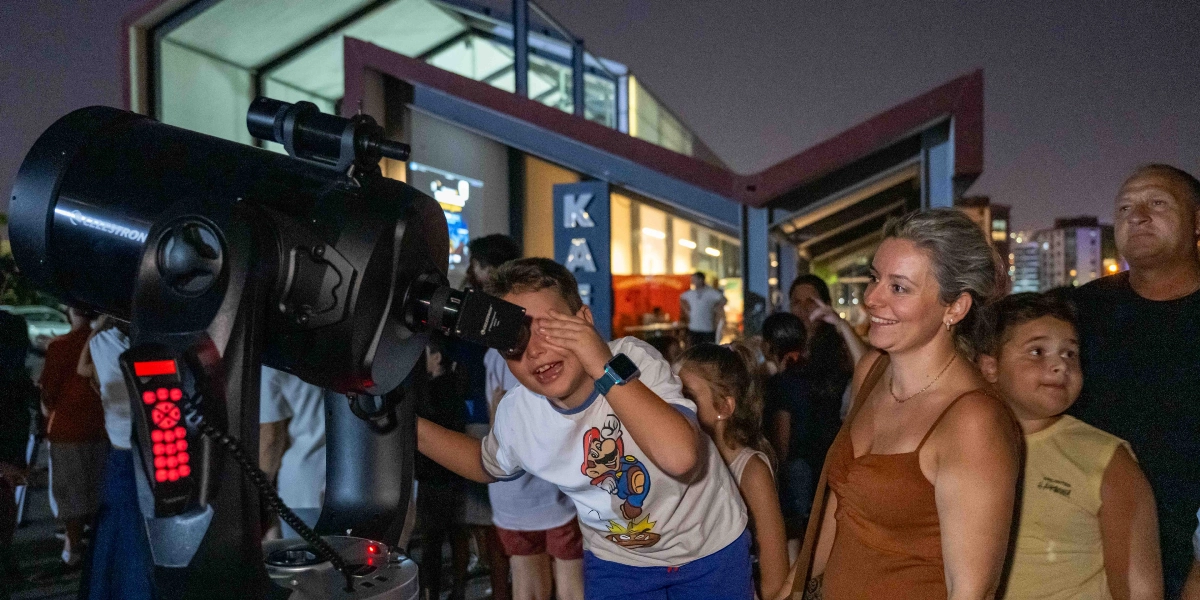 Gece vakti açık havada düzenlenen bir etkinlikte, üzerinde Mario karakteri baskılı beyaz bir tişört giyen küçük bir çocuk, Celestron marka teleskobun merceğine bakıyor. Çocuğun yanında gülümseyerek onu izleyen bir kadın ve başka bir çocuk ile yetişkin bir erkek bulunuyor. Arka planda modern mimariye sahip bir yapı ve kalabalık bir grup yer alıyor.