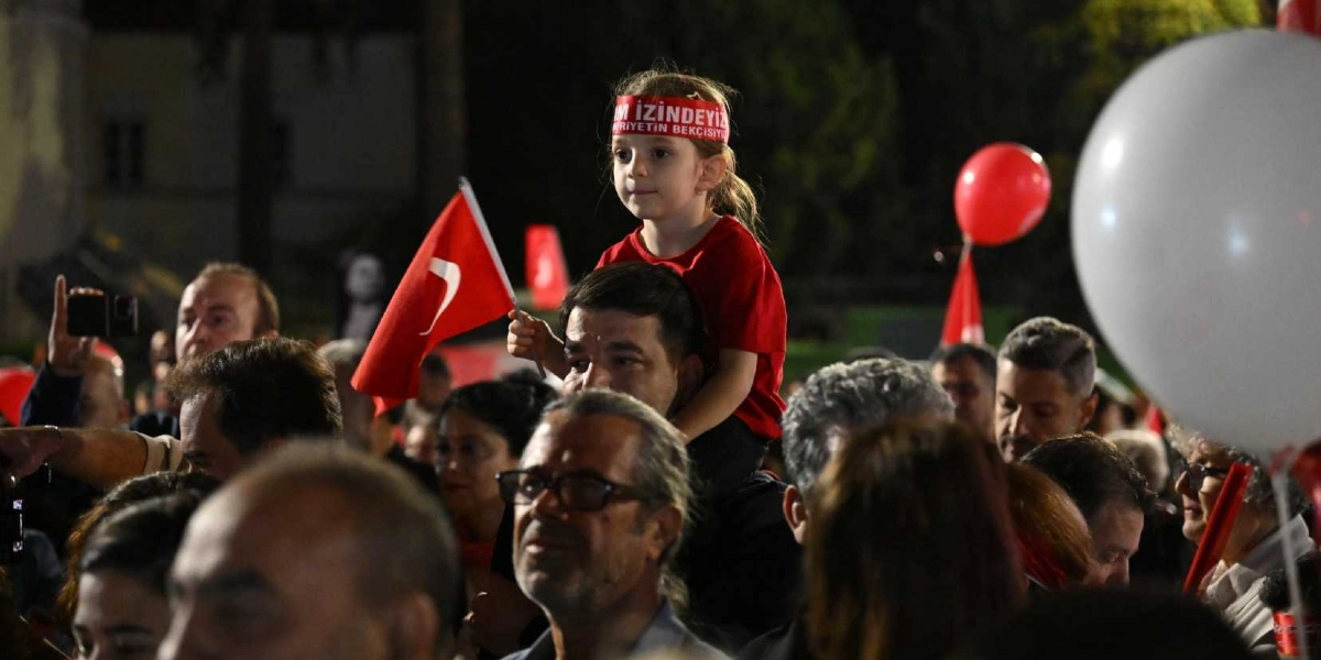 Gece vakti düzenlenen bir Cumhuriyet Bayramı kutlamasında, babasının omuzlarında oturan kırmızı tişörtlü küçük bir kız çocuğu elinde Türk bayrağı tutuyor. Çocuğun başında üzerinde 