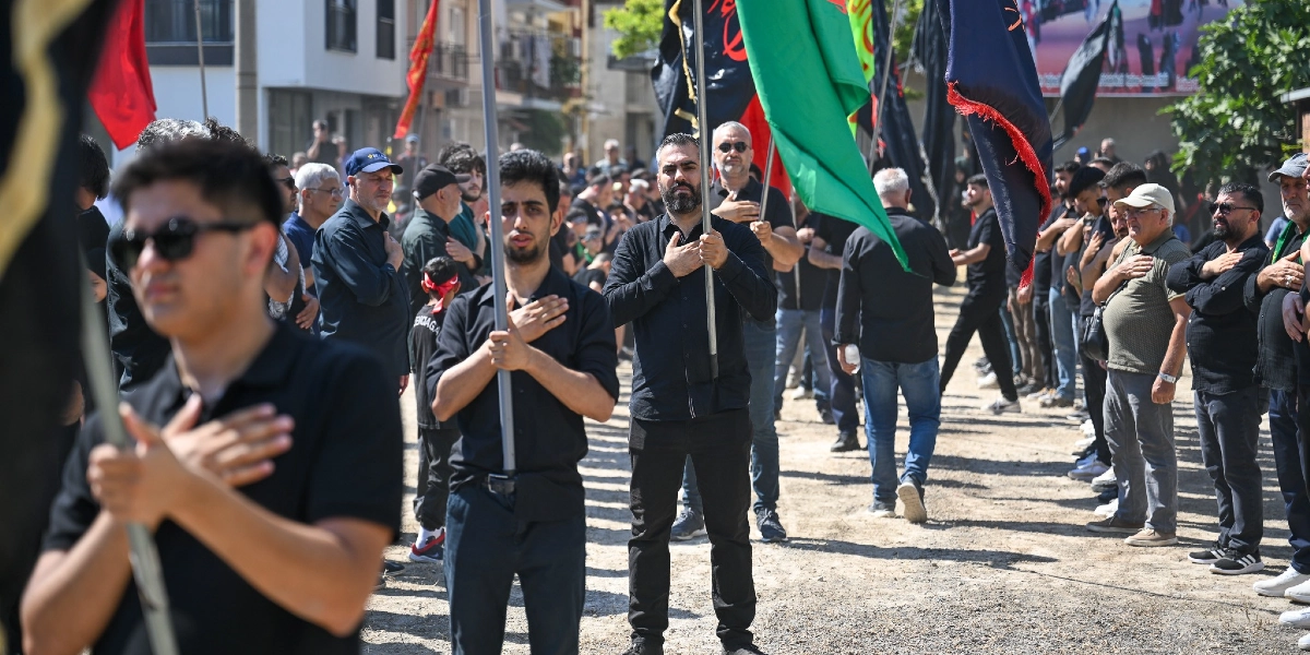 Bornova'da düzenlenen Kerbela anma töreninde, ellerinde siyah, yeşil ve kırmızı flamalar taşıyan, siyah kıyafetler giymiş bir grup erkek, ellerini göğüslerine koyarak yas tutuyor. Toprak bir zemin üzerinde yan yana dizilmiş katılımcılar, ciddiyetle yürüyüşe eşlik ediyor.