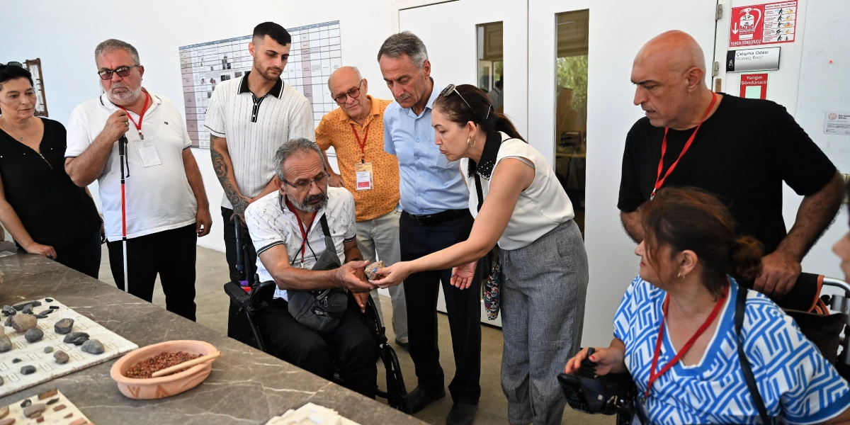 Aydınlık bir müze veya sergi salonunda, tekerlekli sandalyedeki bir katılımcıya rehberlik eden bir kadın, elindeki tarihi bir eseri incelemesi için ona uzatıyor. Etraflarında, boyunlarında yaka kartları asılı olan ve ilgiyle izleyen bir grup katılımcı bulunuyor. Masanın üzerinde çeşitli arkeolojik taş parçaları ve küçük bir kap içinde çakıl taşları sergileniyor. Arka planda duvarda bir çizelge ve yangın güvenliği talimatlarını içeren bir tabela yer alıyor.