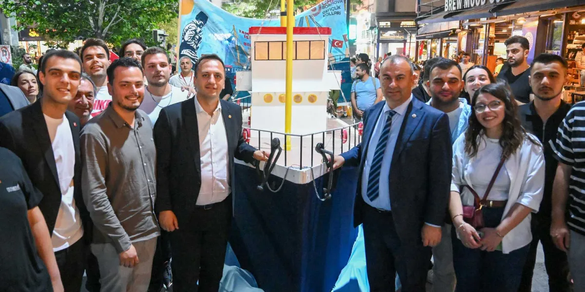 Bornova'da bir sokak etkinliğinde, ortada Bandırma Vapuru'nu temsil eden maket bir geminin etrafında toplanmış, aralarında takım elbiseli siyasetçilerin de bulunduğu genç bir grup insan kameraya gülümseyerek poz veriyor. Arka planda üzerinde Atatürk silüeti ve Türk bayrağı bulunan bir pankart ile çevredeki kafe ve dükkanlar görülüyor.
