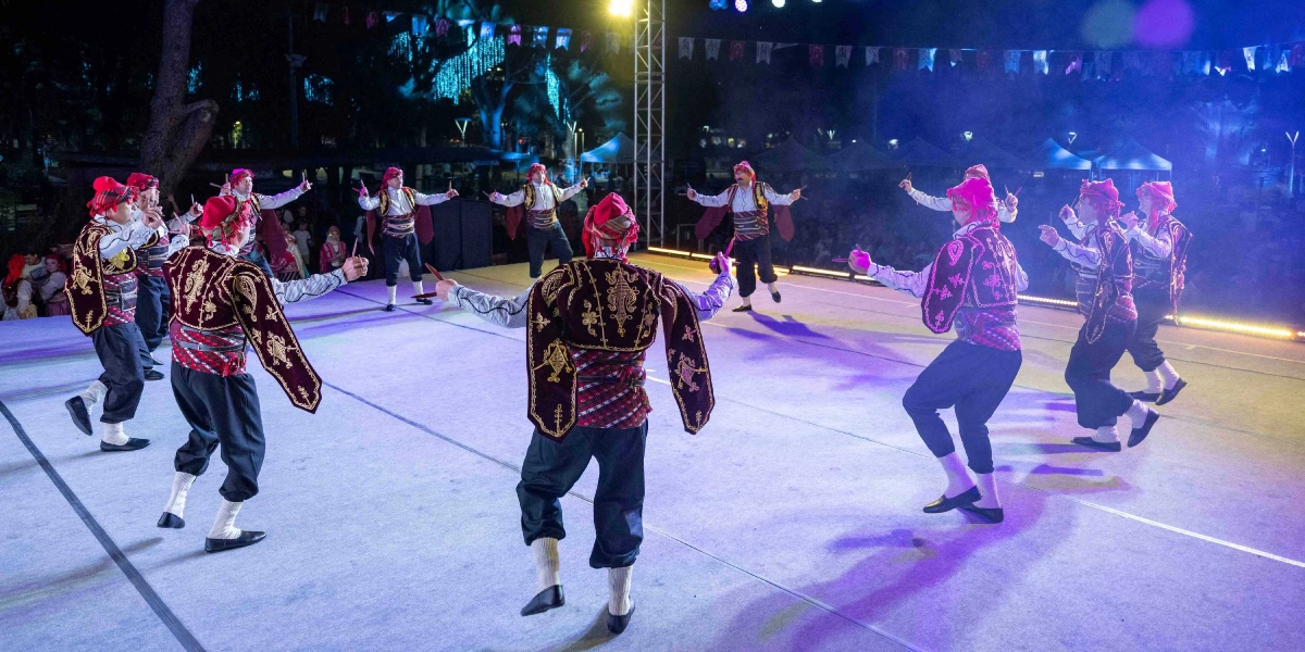 Gece vakti açık hava sahnesinde geleneksel kıyafetler giymiş bir grup erkek dansçı, ellerinde tuttukları kaşıklarla halk oyunu sergiliyor. Dansçılar, üzerlerinde işlemeli koyu renk yelekler, kuşaklar ve başlarında kırmızı feslerle sahne üzerinde dairesel bir formda hareket ediyor. Sahne ışıklarıyla aydınlatılan ortamda, arka planda Türk bayrakları ve seyirciler seçiliyor.