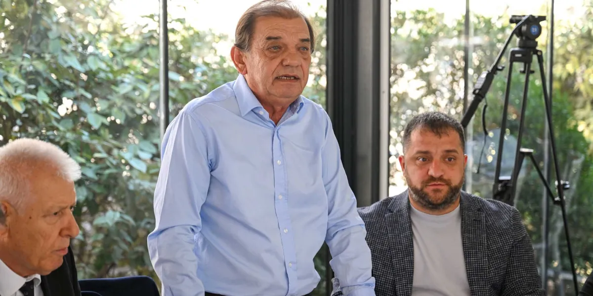 Dramalılar Köşkü'ndeki toplantıda, açık mavi gömlekli orta yaşlı bir erkek ayakta konuşma yaparken, yanında gri ceketli sakallı bir erkek ve sol tarafta takım elbiseli başka bir katılımcı oturuyor. Arka planda camlı bir bölme ve yeşillikler görülürken, sağ tarafta bir tripod üzerine yerleştirilmiş kamera bulunuyor.