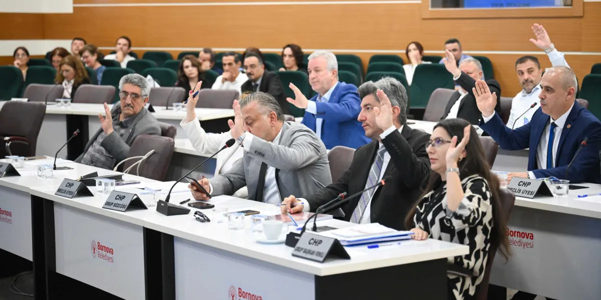 Bornova Belediyesi meclis salonunda düzenlenen toplantıda, masalarda oturan meclis üyeleri bir oylama sırasında ellerini kaldırarak onay veriyor. Ön sırada, üzerinde 
