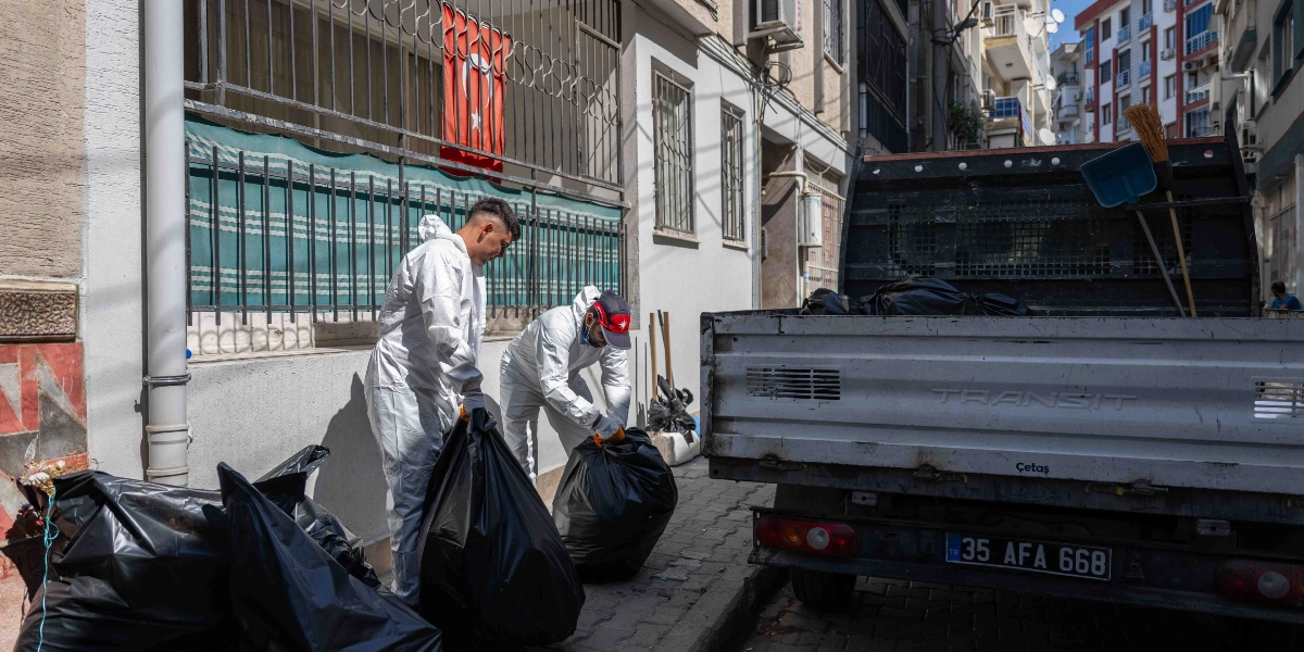 Beyaz koruyucu tulum giymiş iki belediye görevlisi, bir apartmanın önündeki kaldırımda biriktirilmiş çöp poşetlerini toplayarak arkalarında duran kamyonete yüklüyor. Kamyonetin kasası kısmen dolu görünüyor ve arka kısmında süpürge ile faraş bulunuyor. Apartmanın demir parmaklıklarına asılı bir Türk bayrağı dikkat çekiyor.