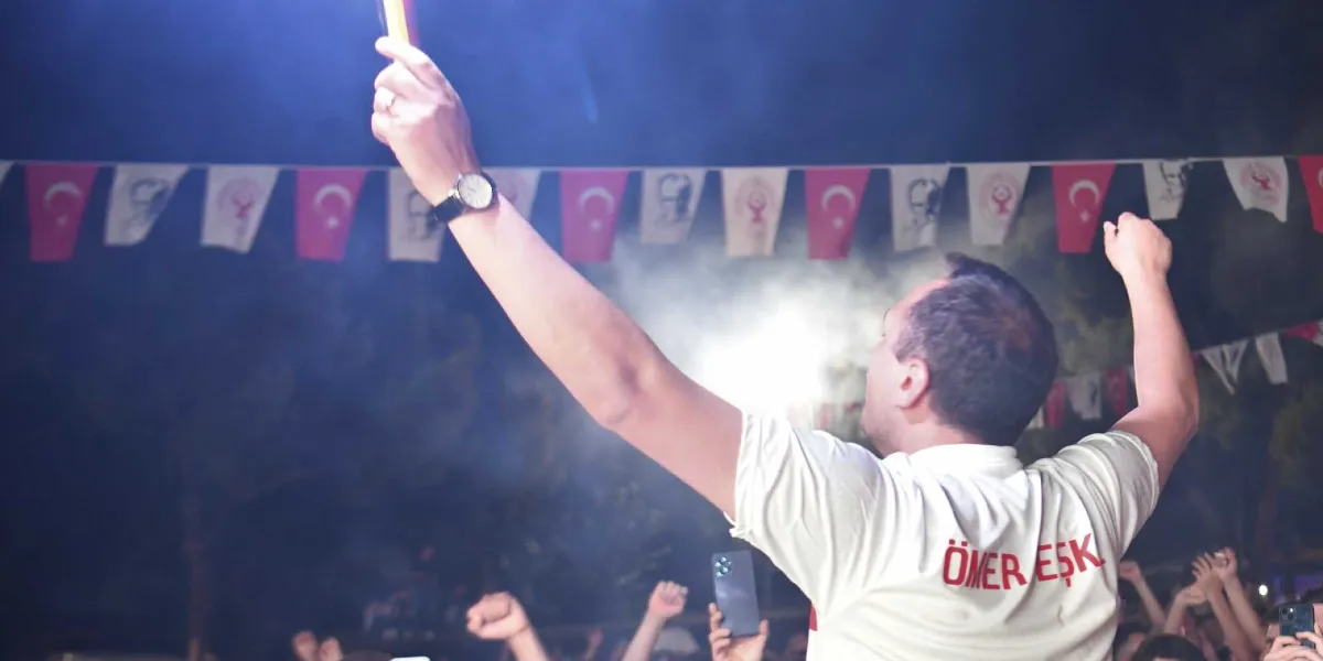 Gece vakti açık havada düzenlenen bir etkinlikte, sırtı kameraya dönük beyaz tişörtlü bir kişi kollarını havaya kaldırmış coşkuyla tezahürat yapıyor. Tişörtünün sırt kısmında kırmızı harflerle ÖMER EŞK yazılı. Kişinin havaya kaldırdığı elinde yanan bir meşale bulunuyor ve etrafa duman yayılıyor. Arka planda ise iplere asılmış Türk bayrakları ve kalabalık bir insan grubu yer alıyor.