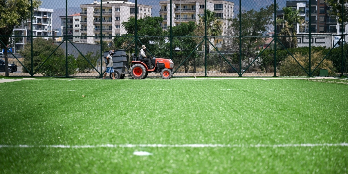 Suni çim kaplı bir futbol sahasında, üzerinde Kubota marka turuncu bir traktör ve ona bağlı bir ekipman bulunan sahada bakım çalışması yapılıyor. Traktörü kullanan bir kişi ve yanında yürüyen başka bir görevli, sahanın tel örgüyle çevrili kenar kısmında ilerliyor. Arka planda ise çok katlı apartmanlar ve ağaçlar görünüyor.