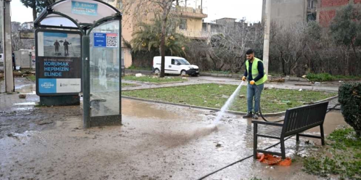 Bornova'da sel sonrası çamurla kaplanan bir otobüs durağı çevresinde, üzerinde sarı ikaz yeleği bulunan bir belediye çalışanı hortumla tazyikli su sıkarak temizlik yapıyor. Durağın yanında çamurlu bir zemin, arka planda ise park halindeki beyaz bir araç ve ağaçlar görülüyor. Durak üzerindeki reklam panosunda 