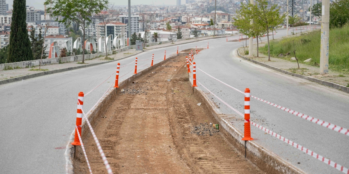 Bornova'da asfaltı kazılmış ve yol çalışması devam eden bir varyant yolu. Yolun ortasındaki kazı alanı, güvenlik amacıyla turuncu beyaz plastik dubalar ve şeritlerle çevrilmiş durumda. Arka planda ise şehir yerleşimini gösteren binalar ve ağaçlar yer alıyor.