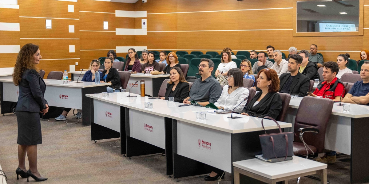 Bornova Belediyesi'ne ait ahşap panellerle kaplı bir konferans salonunda, ayakta duran bir kadın konuşmacı, sıralarda oturan katılımcılara hitap ediyor. Katılımcılar, üzerinde Bornova Belediyesi logosu bulunan beyaz masalarda oturarak sunumu dinliyor. Salonun arka duvarında, üzerinde 