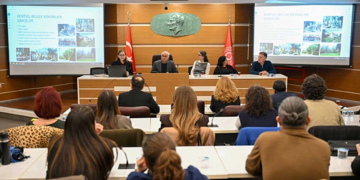 Bornova'nın tarihi kimliğine yönelik bir toplantıda, bir masanın etrafında oturan beş kişilik bir konuşmacı grubu ve onları dinleyen katılımcılar yer alıyor. Salonun ön kısmındaki iki büyük ekranda 