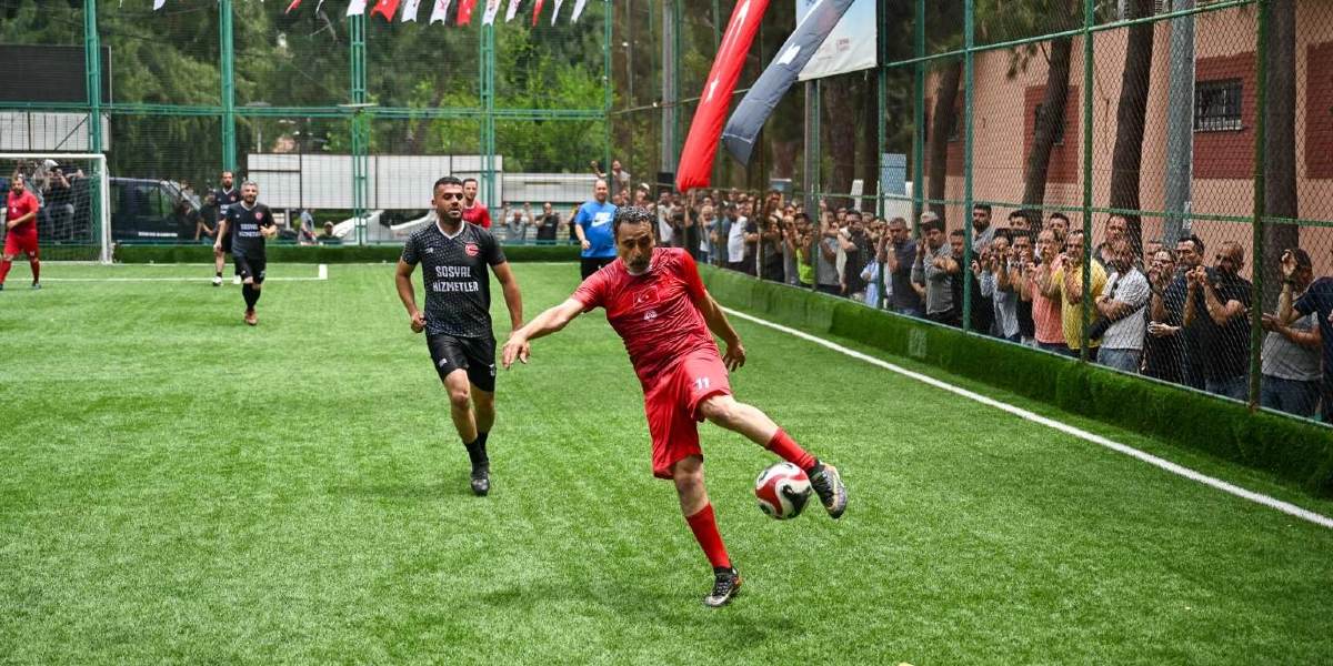 Halı sahada oynanan bir futbol maçında, kırmızı formasıyla topa vuran bir oyuncu ve onu takip eden siyah formalı rakip oyuncu görülüyor. Saha kenarındaki tel örgülerin arkasında maçı izleyen bir kalabalık yer alıyor ve sahada Türk bayrakları asılı duruyor.