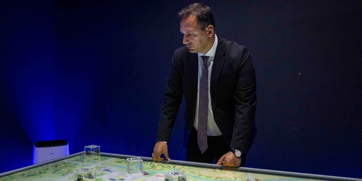 Koyu mavi bir arka plan önünde, beyaz gömlek ve lacivert kravatlı, siyah takım elbiseli bir erkek, üzerinde Bornova'nın şehir planını temsil eden dijital bir haritanın bulunduğu ışıklı bir masayı dikkatle inceliyor. Masanın üzerinde, artırılmış gerçeklik veya etkileşimli bir sistemin parçası gibi görünen, üzerinde kare kod benzeri işaretler bulunan şeffaf küpler yer alıyor.