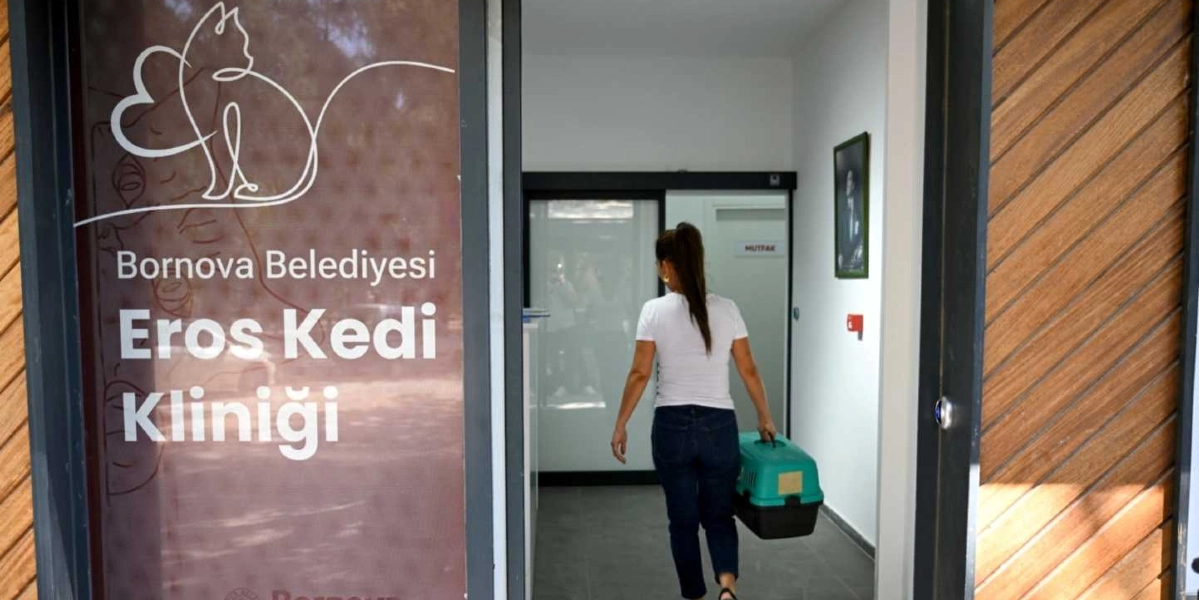 Bornova Belediyesi Eros Kedi Kliniği'nin girişinde, elinde yeşil bir kedi taşıma çantasıyla içeri giren beyaz tişörtlü ve kot pantolonlu bir kadın görülüyor. Kliniğin cam kapısında beyaz çizgilerle çizilmiş bir kedi silüeti ve 