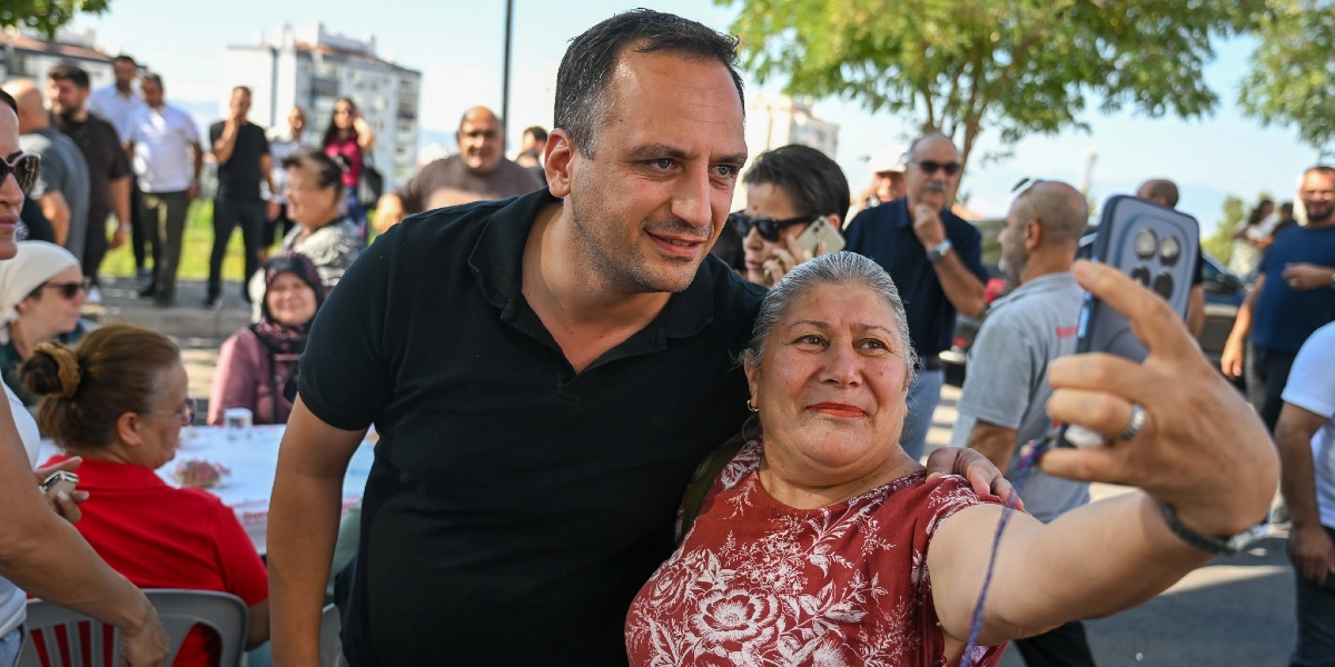 Siyah polo yaka tişörtlü bir erkek, açık havada düzenlenen bir etkinlikte çiçek desenli kiremit rengi bluz giyen bir kadınla yan yana durarak selfie çekiliyor. Arka planda Kent Bostanı'ndaki etkinlik alanında oturan ve ayakta duran diğer katılımcılar ile ağaçlar görülüyor.