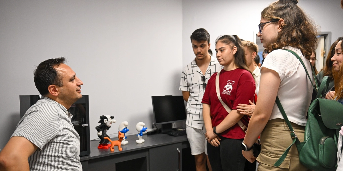Aydınlık bir ofis ortamında, çizgili gömlek giyen bir eğitmen, karşısında duran ve dikkatle dinleyen gençlerden oluşan bir gruba 3D yazıcıdan çıkmış küçük figürleri gösteriyor. Masanın üzerinde Gargamel, Şirinler ve turuncu bir kedi figürü bulunuyor. Öğrenciler arasında bordo tişörtlü bir genç kız ve beyaz tişörtlü, gözlüklü bir başka genç kız ön planda yer alıyor. Arka planda ise beyaz çizgili gömlek giyen bir genç erkek ve diğer öğrenciler bulunuyor.