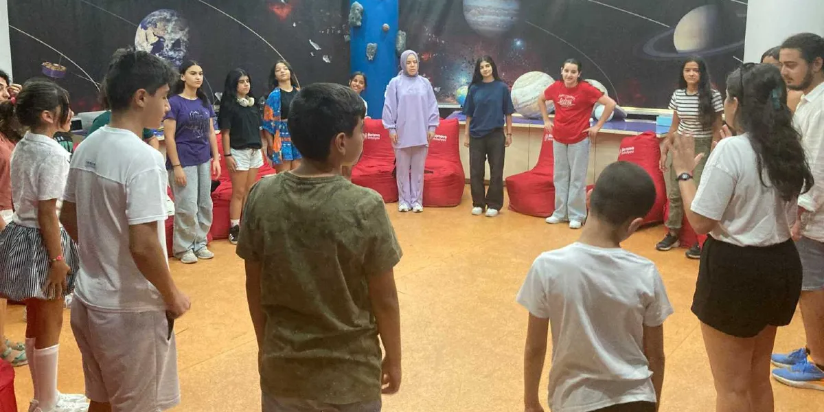 Güneş sistemi temalı bir duvar görselinin önünde, geniş bir daire oluşturmuş şekilde duran bir grup çocuk ve genç. Bornova Belediyesi logolu kırmızı armut koltukların bulunduğu sınıfta, bir eğitmen eşliğinde etkileşimli bir etkinlik gerçekleştiren katılımcılar birbirlerine dönük şekilde duruyor.