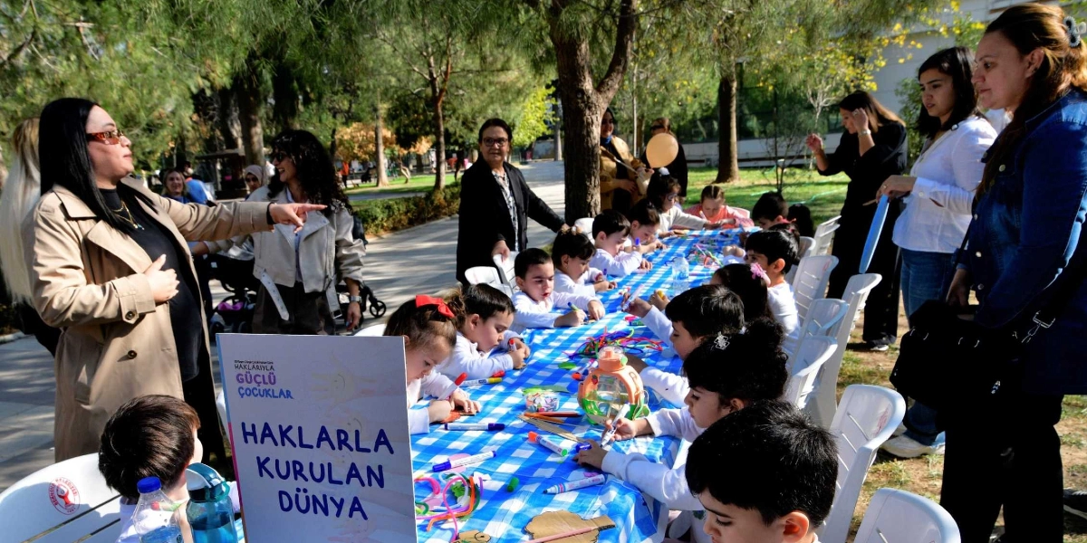 Açık havada, ağaçlık bir alanda uzun bir masanın etrafında toplanmış çocuklar resim ve el işi etkinlikleri yapıyor. Mavi beyaz kareli örtüyle kaplı masada çocuklar boya kalemleri ve kağıtlarla ilgilenirken, yanlarında duran yetişkinler onlara eşlik ediyor. Masanın ön kısmında üzerinde 