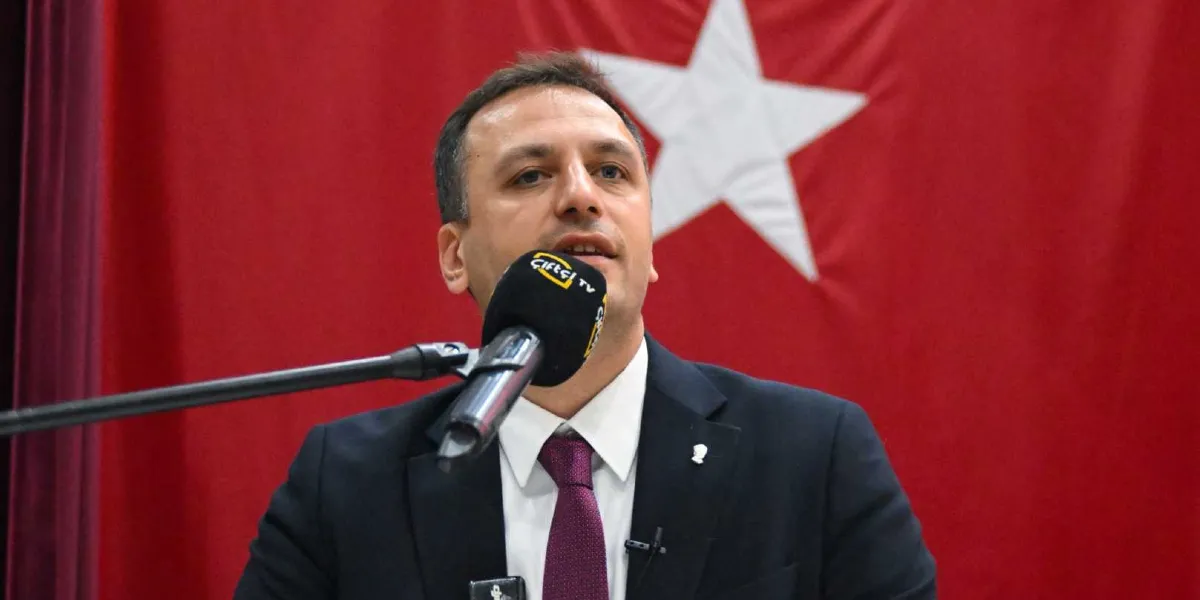 Koyu renk takım elbise ve bordo kravat giymiş bir erkek, arkasında kırmızı zemin üzerine beyaz yıldız bulunan bir bayrakla, üzerinde Çiftçi TV logosu bulunan bir mikrofona konuşma yapıyor.