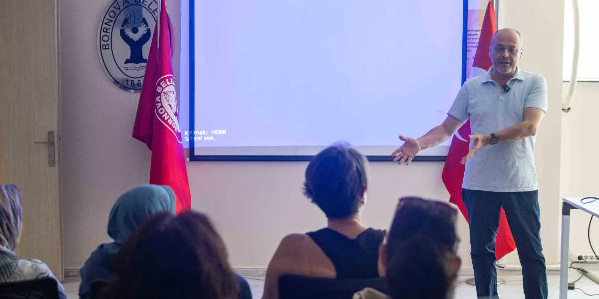 Bornova Belediyesi logosunun ve Türk bayrağının yer aldığı bir salonda, gözlüklü ve açık renkli polo yaka tişört giyen bir konuşmacı, arkasındaki projeksiyon ekranına karşı el hareketleriyle bir gruba sunum yapıyor.