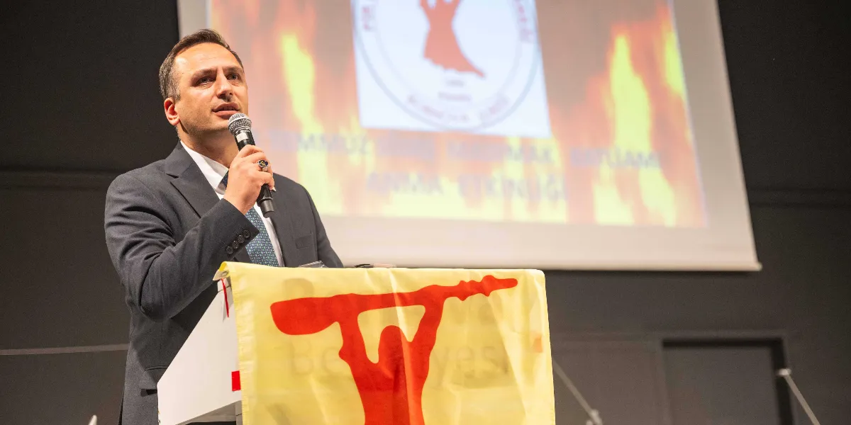 Koyu renk takım elbiseli bir erkek, kürsüde mikrofonla konuşma yapıyor. Kürsünün önünde üzerinde kırmızı renkli semah dönen insan figürü bulunan sarı bir örtü serili. Arka planda yer alan projeksiyon perdesinde ise alev temalı bir arka plan üzerinde aynı semah figürü ve 