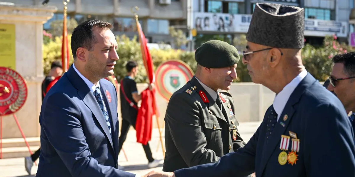 Bornova Belediye Başkanı Mustafa İduğ, Gaziler Günü töreninde resmi üniformalı bir askeri yetkili ve madalyalı bir gazi ile tokalaşarak selamlaşıyor. Arka planda tören alanında bayraklar ve çelenkler yer alıyor.