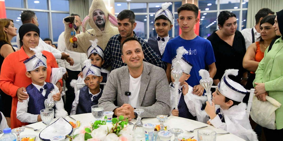Sünnet kıyafetleri giymiş çocukların ve ailelerinin katıldığı bir etkinlikte, masanın merkezinde oturan bir adam ve çevresindeki grup gülümsüyor. Sünnet kıyafeti içindeki çocuklar ellerinde asalarla masada otururken, arkalarında tavşan kostümlü bir animatör ve yetişkinler yer alıyor. Masada yemek tabakları ve içecekler bulunuyor.