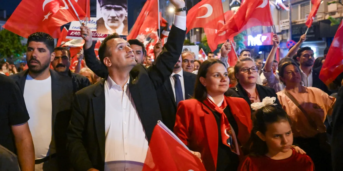 Akşam saatlerinde kalabalık bir grup, ellerinde Türk bayrakları ve Mustafa Kemal Atatürk’ün portresini taşıyarak bir yürüyüş gerçekleştiriyor. Ön planda, siyah ceketli ve beyaz gömlekli bir erkek elini havaya kaldırmış, yanında kırmızı ceketli bir kadın ve diğer katılımcılarla birlikte coşkulu bir şekilde ilerliyor. Arka planda ise şehir ışıkları ve yürüyüşe katılan çok sayıda insan yer alıyor.