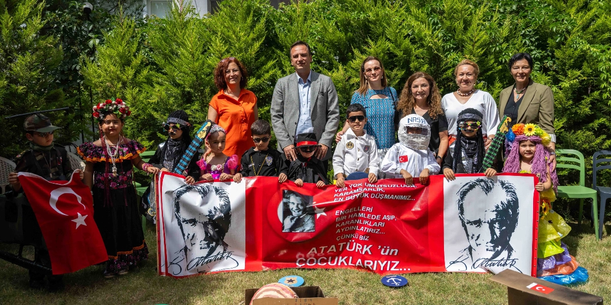 Yeşil bir bahçede, farklı kostümler giymiş çocukların ve yetişkinlerin bir arada olduğu bir kutlama etkinliği. Çocuklar, üzerinde Atatürk portreleri ve 