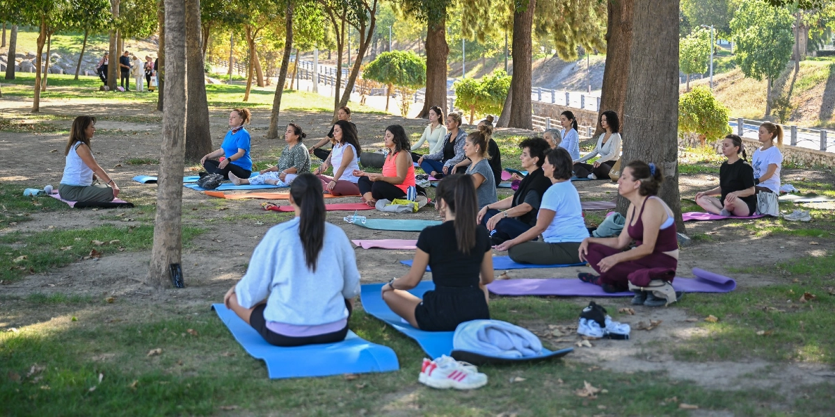 Ağaçlık bir park alanında, yoga matları üzerinde bağdaş kurarak oturan ve meditasyon yapan bir grup kadın. Kadınlar farklı renklerde spor kıyafetleri giymiş, huzurlu bir ortamda bir araya gelmişler. Arka planda ağaçlar ve hafif eğimli bir arazi görülüyor.