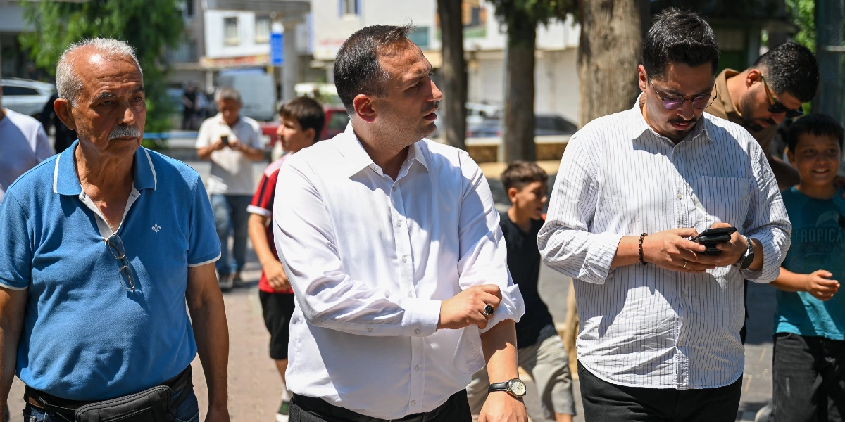 Açık havada, güneşli bir günde yan yana yürüyen üç erkekten soldaki mavi polo yaka tişörtlü ve bıyıklı, ortadaki beyaz gömlekli ve sağdaki çizgili gömlekli olan ise elindeki telefona bakıyor. Arka planda ise yürüyen çocuklar ve diğer insanlar yer alıyor.