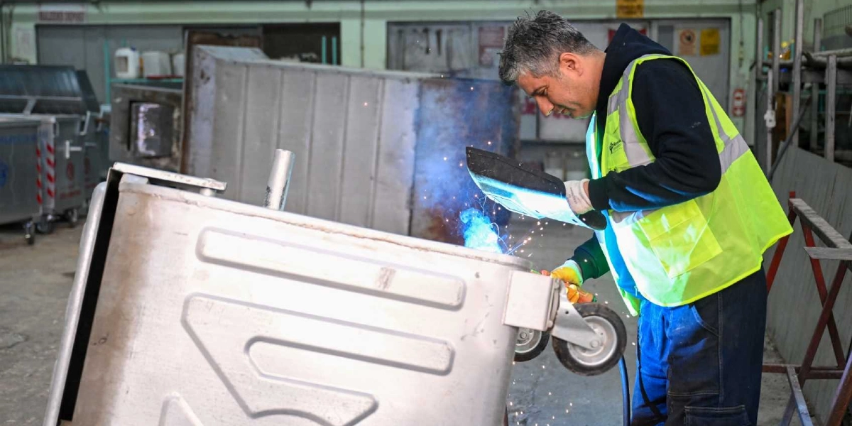 Atölye ortamında, üzerinde Bornova Belediyesi logosu bulunan sarı reflektörlü iş yeleği giymiş bir işçi, metal bir çöp konteynerine kaynak yapıyor. İşçi, koruyucu maskesini tutarak metal parçaları birleştirirken etrafa kıvılcımlar saçılıyor. Arka planda ise üretimi tamamlanmış veya tamir edilmeyi bekleyen diğer metal çöp konteynerleri görülüyor.