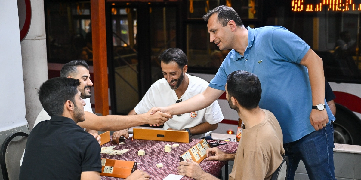 Mavi polo yaka tişörtlü bir erkek, okey masasında oturan genç bir adamla el sıkışıyor. Masanın etrafında oturan diğer üç genç adam, oyun oynamaya devam ederken gülümseyerek bu anı izliyor. Ortamda okey taşları ve ıstakalar bulunuyor.