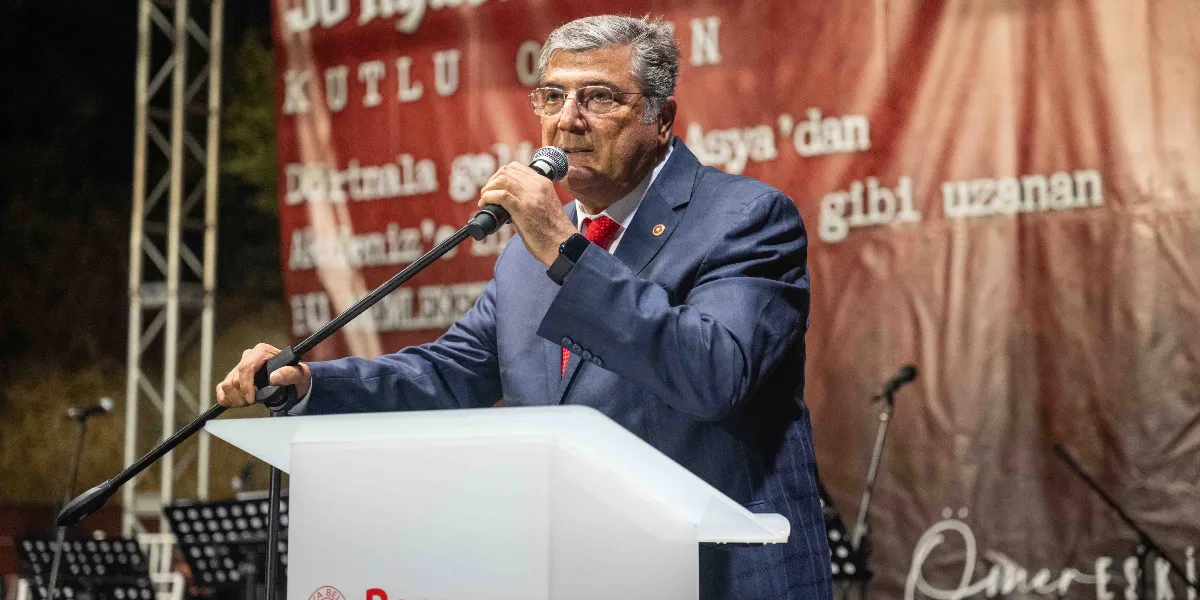 Koyu renk takım elbiseli, kırmızı kravatlı ve gözlüklü bir erkek, üzerinde Bornova Belediyesi logosu bulunan beyaz bir kürsüde mikrofonla konuşma yapıyor. Arkasındaki kırmızı renkli afişte Nazım Hikmet’in 