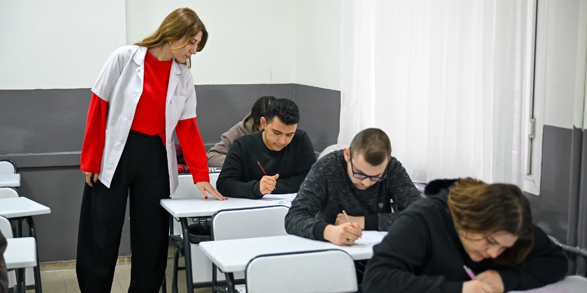 Sınıf ortamında, beyaz önlük giymiş bir eğitmen, sıralarda oturup ders çalışan öğrencilerin çalışmalarını kontrol ediyor. Öğrenciler masalarında oturmuş, dikkatle not alıyor veya yazı yazıyor.