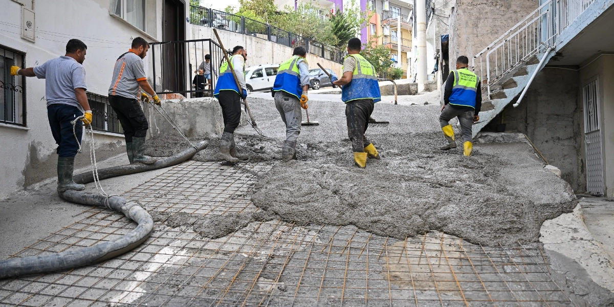 Bornova Belediyesi işçileri, eğimli bir sokakta beton dökme ve yol yenileme çalışması yapıyor. Üzerlerinde reflektörlü yelekler ve çizmeler bulunan altı işçi, hortumla beton yayıyor, tırmıklarla düzeltme yapıyor ve demir donatıların üzerine beton döküyor.