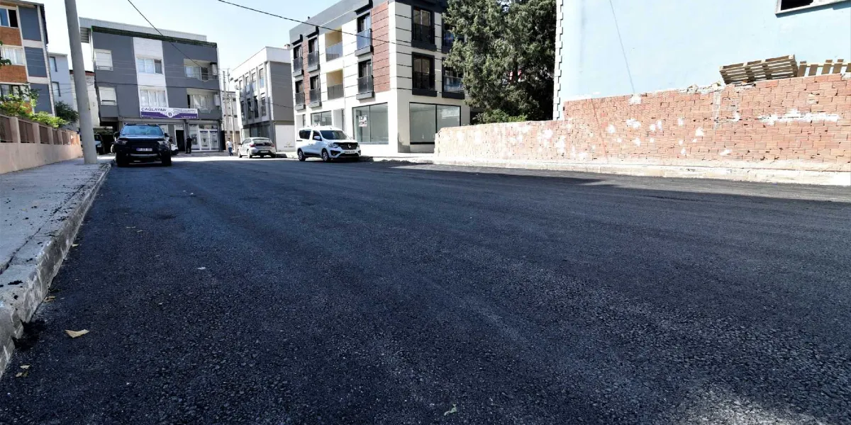Bornova'da yeni asfaltlanmış, pürüzsüz ve siyah renkteki geniş bir cadde. Yolun kenarlarında modern apartmanlar, park halindeki araçlar ve sağ tarafta alçak bir tuğla duvar bulunuyor.