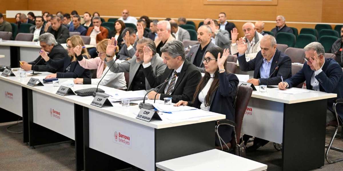 Bornova Belediyesi meclis salonunda düzenlenen toplantıda, katılımcıların bir oylama sırasında ellerini kaldırarak onay verdikleri görülüyor. Masaların üzerinde Bornova Belediyesi logoları ve CHP grup temsilcilerinin isimlikleri yer alıyor. Toplantı salonunda çok sayıda kişi sıralı masalarda oturuyor ve önlerinde evraklar bulunuyor.