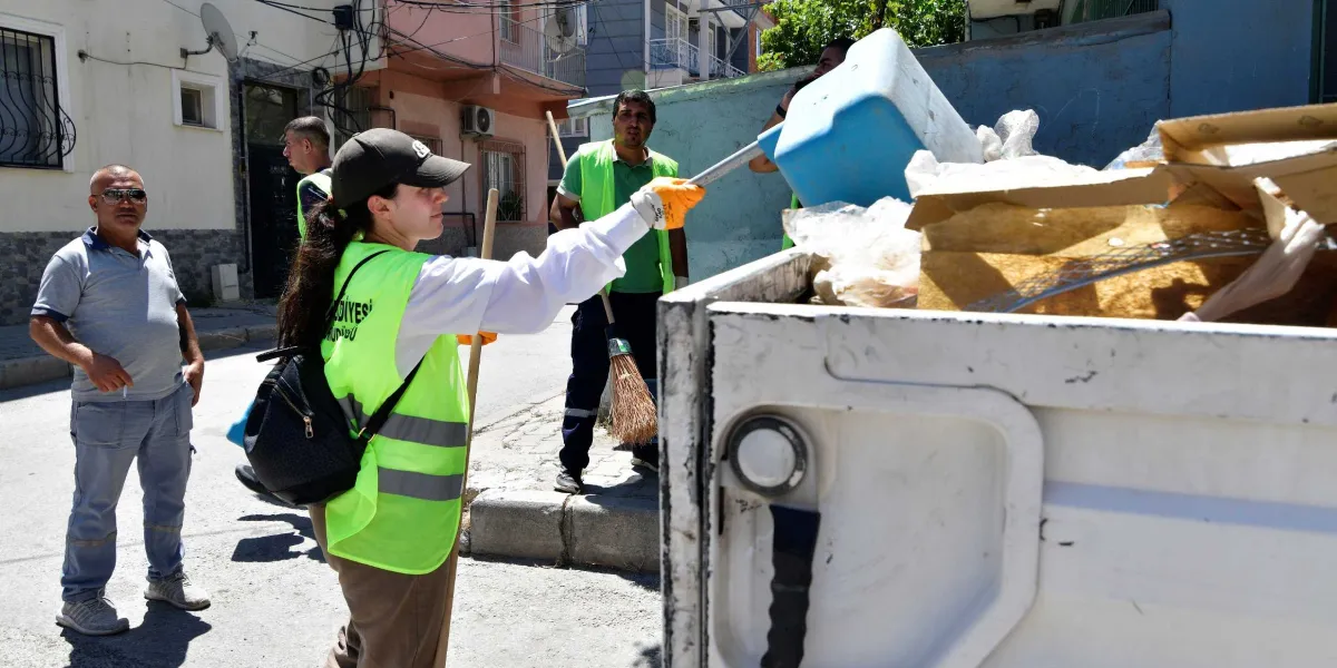 Bornova Belediyesi bünyesinde çalışan temizlik görevlileri, sokakta çöp toplama işlemi yapıyor. Sarı fosforlu yelek giyen bir kadın görevli, elindeki mavi çöp küreğiyle atıkları kamyonun kasasına boşaltırken, yanında süpürgesiyle bekleyen bir erkek çalışma arkadaşı ve sivil giyimli bir vatandaş bulunuyor.