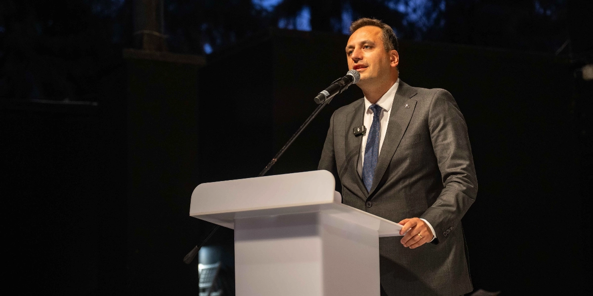 Koyu renkli bir arka plan önünde, beyaz bir kürsü arkasında duran ve mikrofonla konuşma yapan, gri takım elbiseli ve mavi kravatlı bir erkek.