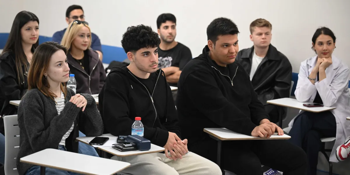 Bir sınıfta sıralarda oturan ve dikkatle dinleyen üniversite öğrencileri. Ön planda siyah kapüşonlu sweatshirt giymiş genç bir erkek ve yanında koyu renkli hırkalı bir kadın bulunuyor; arka sıralarda ise farklı kıyafetler içindeki diğer öğrenciler yer alıyor. Öğrencilerin önündeki sıraların üzerinde su şişeleri, telefonlar ve kişisel eşyalar duruyor.