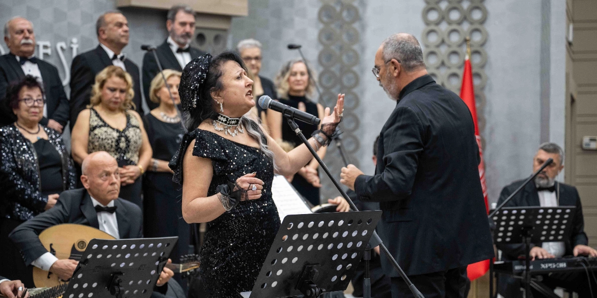 Siyah payetli bir elbise giymiş kadın solist, mikrofon başında şarkı söylerken, yanında orkestra şefi ve arkasında koro üyeleri yer alıyor. Müzisyenler, ud ve klavye gibi enstrümanlar eşliğinde sahne alıyor. Arka planda Türk bayrağı ve dekoratif duvar panelleri bulunuyor.